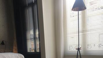 Um quarto com cama, sofá escuro, almofada estampada e luminária de chão perto de uma janela com persianas.
