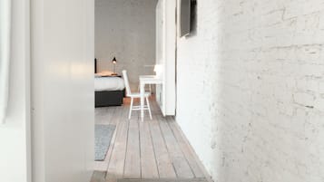 Un couloir étroit avec un plancher en bois, des murs blancs et une chambre visible, où l’on aperçoit un lit et une chaise.