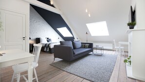 Een loft-kamer met schuine wanden, een grijze bank, een witte eettafel en een kleine witte stoel.