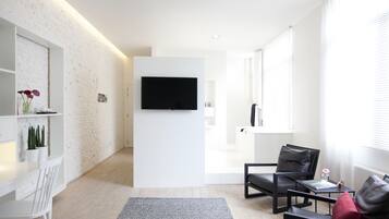 Un salon moderne avec un téléviseur à écran plat fixé à un mur blanc, un canapé en cuir noir, une table à manger blanche avec des chaises, et une petite table d’appoint sur laquelle se trouvent un livre et une tasse.