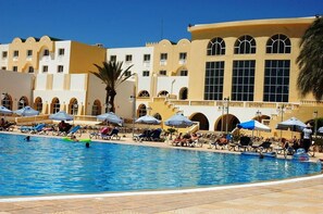 A swimming pool with lounge chairs and umbrellas, surrounded by a building with arches and large windows. - Djerba Castille Hotel (Aghir)