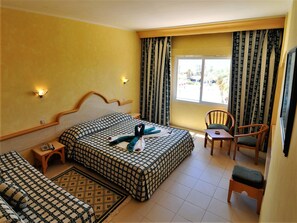 Ein Hotelzimmer mit kariertem Bett, einem kleinen Tisch, einem Stuhl und Blick ins Freie durch ein Fenster.