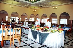 A banquet hall with multiple tables set for dining, a central buffet area, and a kitchen in the background. - Djerba Castille Hotel (Aghir)