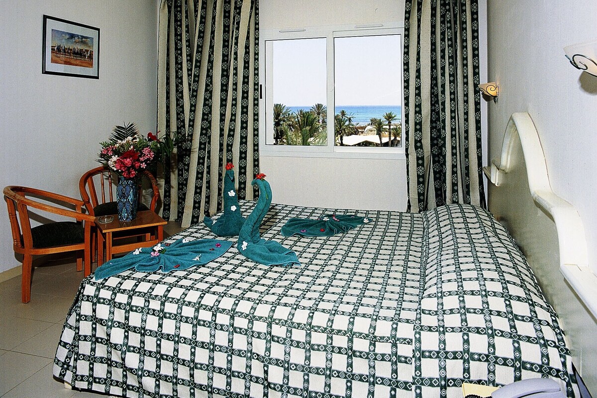A bedroom with a bed, a chair, a table with a vase of flowers, and curtains.
