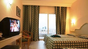 Habitación de hotel con televisión, cama con ropa de cama estampada, una silla y un balcón con cortinas.