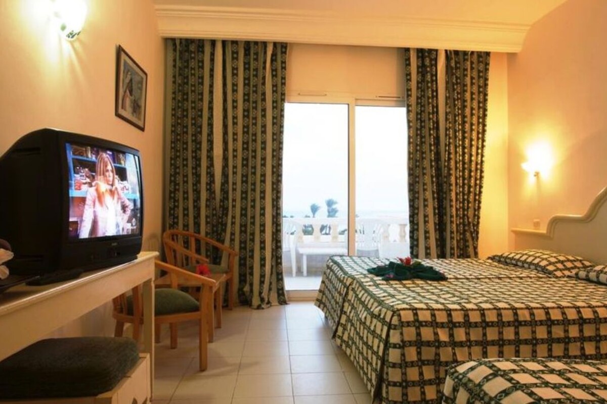 A hotel room with a television, a bed with patterned bedding, a chair, and a balcony with curtains.