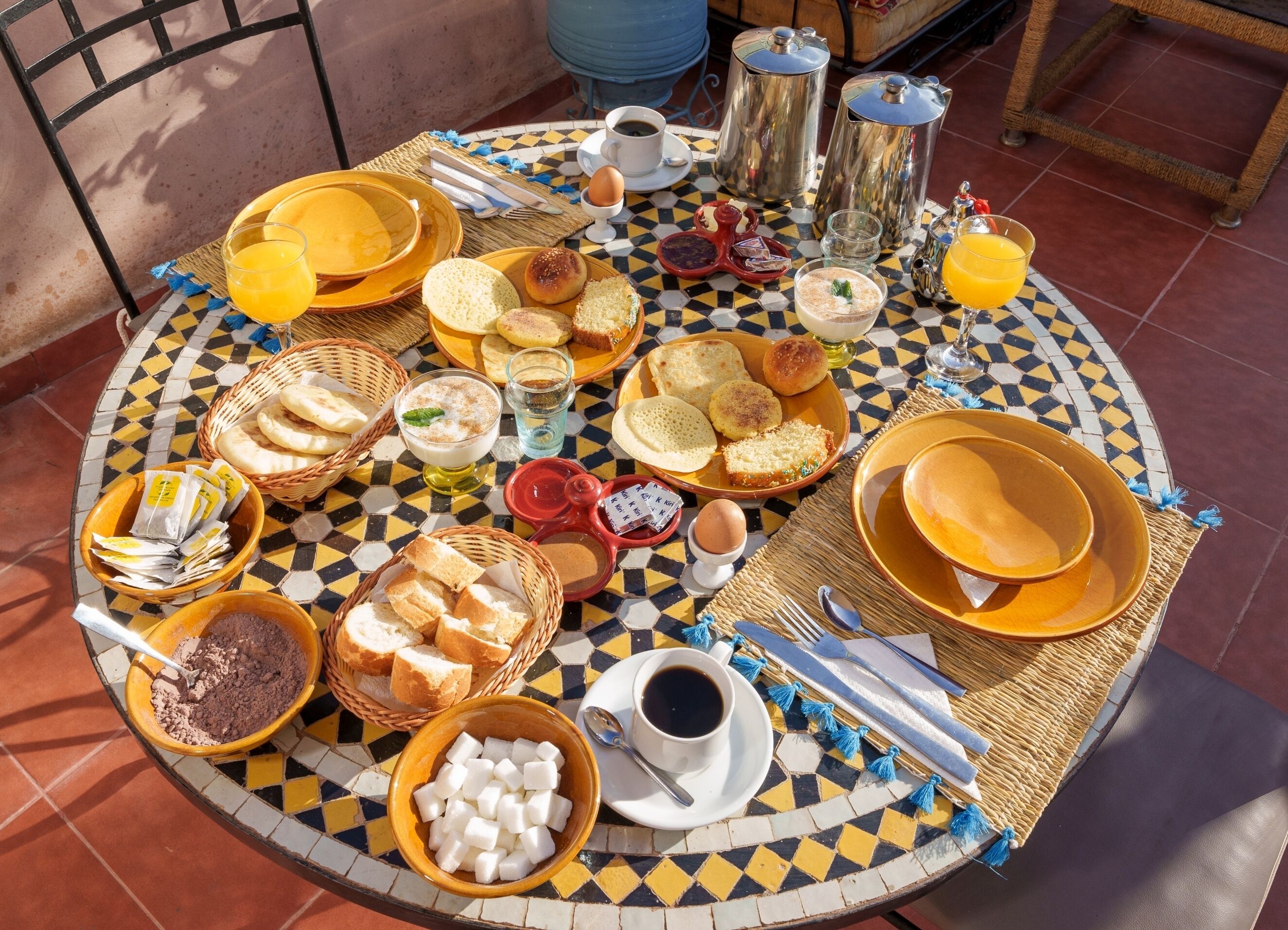 Una mesa redonda con un estampado de baldosas de cuadros, llena de diversos alimentos para el desayuno como pan, pasteles y bebidas, evocando un ambiente para disfrutar de un desayuno tranquilo.