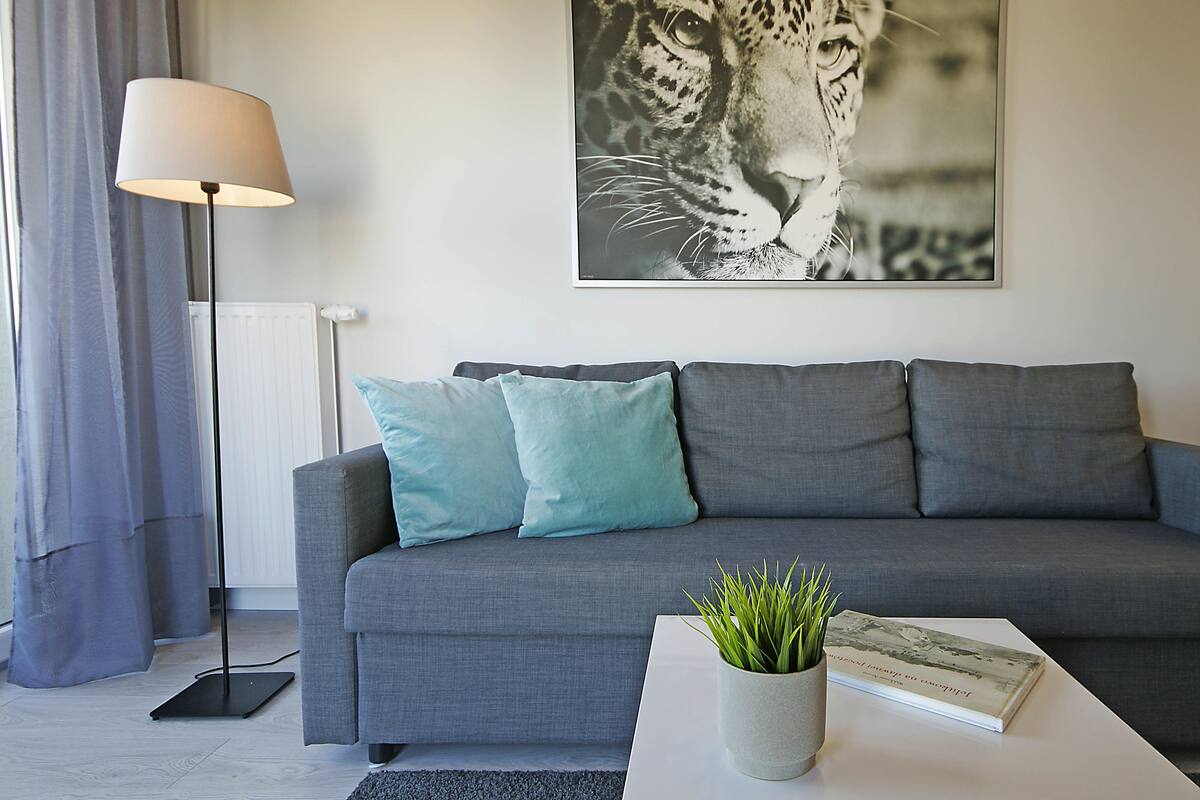 A modern living room with a grey sofa, teal cushions, a floor lamp, a white coffee table with a plant and a book, and a leopard print artwork on the wall.