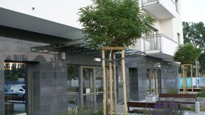 A modern building with a glass entrance, a wooden tree structure, and benches. - IRS ROYAL APARTMENTS - IRS Albatros (Gdansk)
