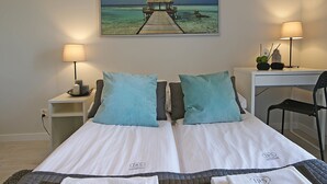A hotel room with a bed, two pillows, a desk, a chair, a lamp, a painting of a beach, and towels with the hotel logo. - IRS ROYAL APARTMENTS - IRS Albatros (Gdansk)