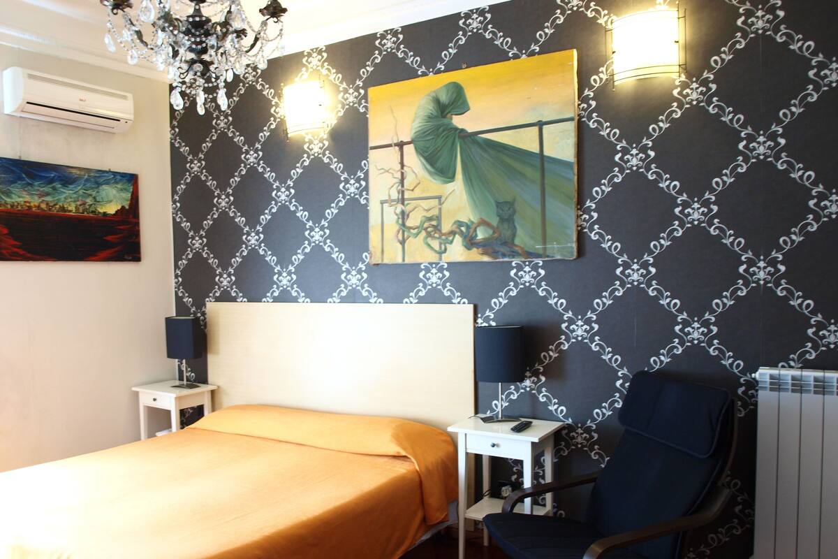 A hotel room with a bed, a chair, a desk, a lamp, an air conditioner, and a painting on the wall.