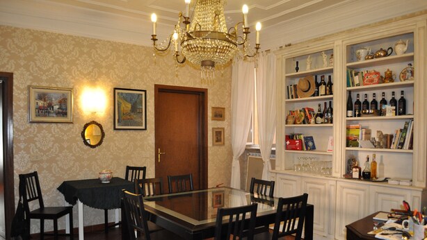 A dining room with a glass-top table, wooden chairs, a chandelier, and a bookshelf filled with books and bottles. - B&B Santa Bibiana (Rome)