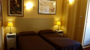 A hotel room with two single beds, a nightstand with a lamp, a wall-mounted painting, and a door with a light switch. - B&B Santa Bibiana (Rome)