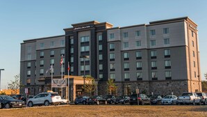 A multi-story hotel with a 'NOW OPEN' sign, a parking lot with various vehicles, and a clear sky. - Hampton Inn & Suites by Hilton Bolton (Caledon)