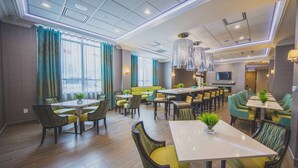 A modern dining area with multiple tables and chairs, a sofa, and a TV screen. - Hampton Inn & Suites by Hilton Bolton (Caledon)