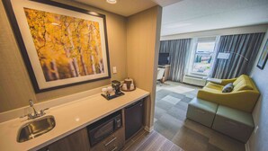 A modern kitchen with a sink, microwave, and coffee maker, a framed artwork above the counter, and a sofa with a view of the outside. - Hampton Inn & Suites by Hilton Bolton (Caledon)
