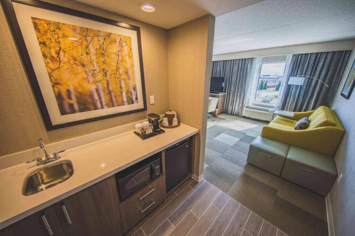 A compact hotel room with a kitchenette, a sofa, a large window, and a framed artwork on the wall.
