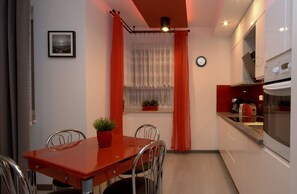A compact kitchen with a dining area, featuring a red table, two chairs, a potted plant, and a window with curtains. - IRS ROYAL APARTMENTS - IRS Aviator (Gdansk)