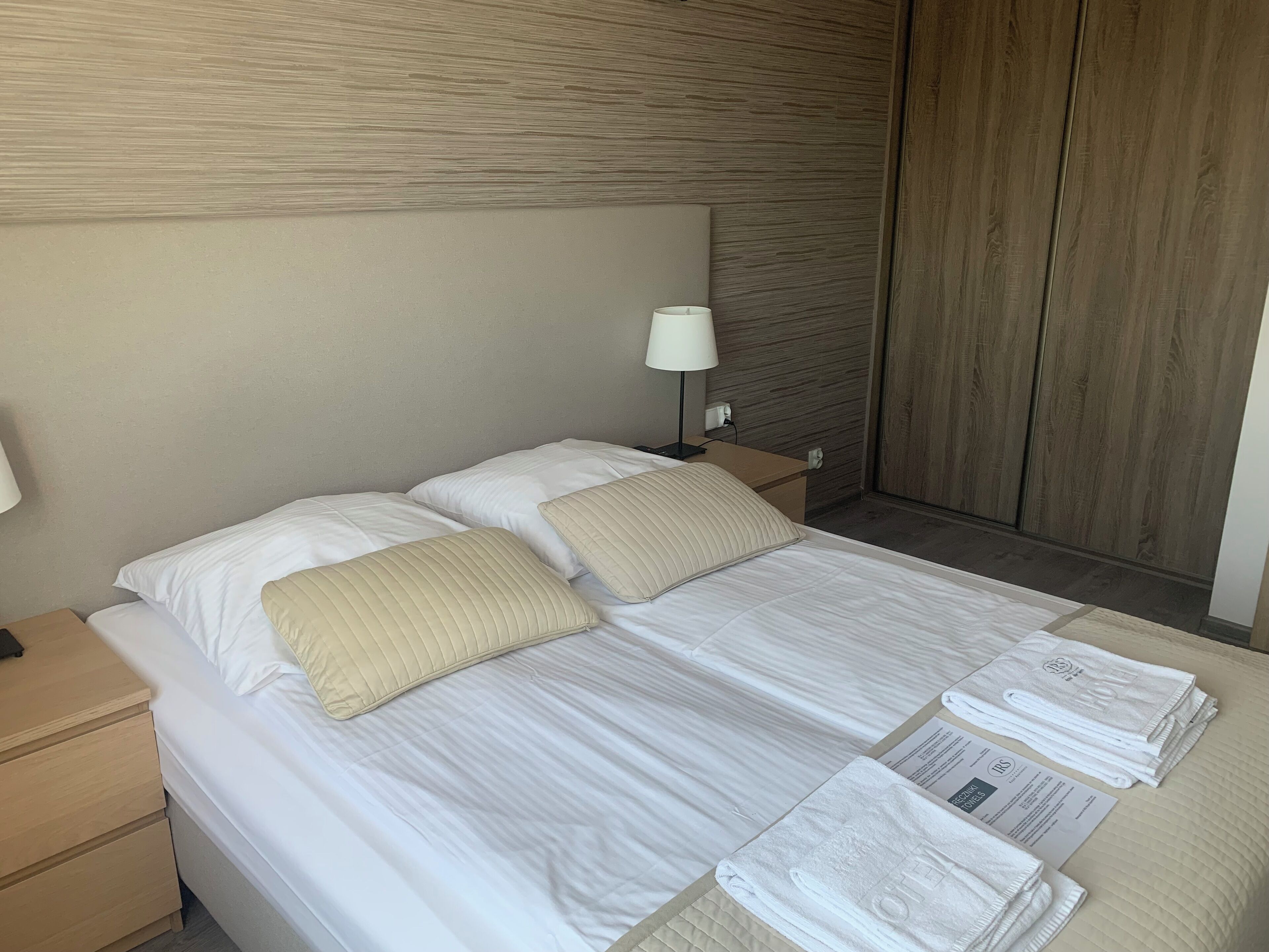 Una cama bien hecha con sábanas blancas y almohadas color beige, una mesita de noche con lámpara y un armario de madera en una habitación de hotel.