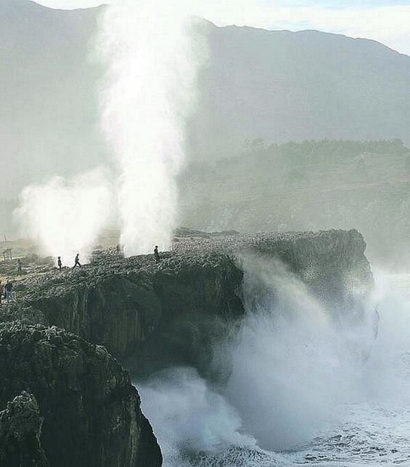 A coastal landscape with a prominent cliff and a geyser emitting steam.