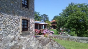 A stone building with a window and a balcony, surrounded by a stone wall and a grassy area with flowers. - Hotel Rural Ovio (Llanes)