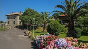 A stone building with a paved driveway, surrounded by lush greenery and flowering plants. - Hotel Rural Ovio (Llanes)