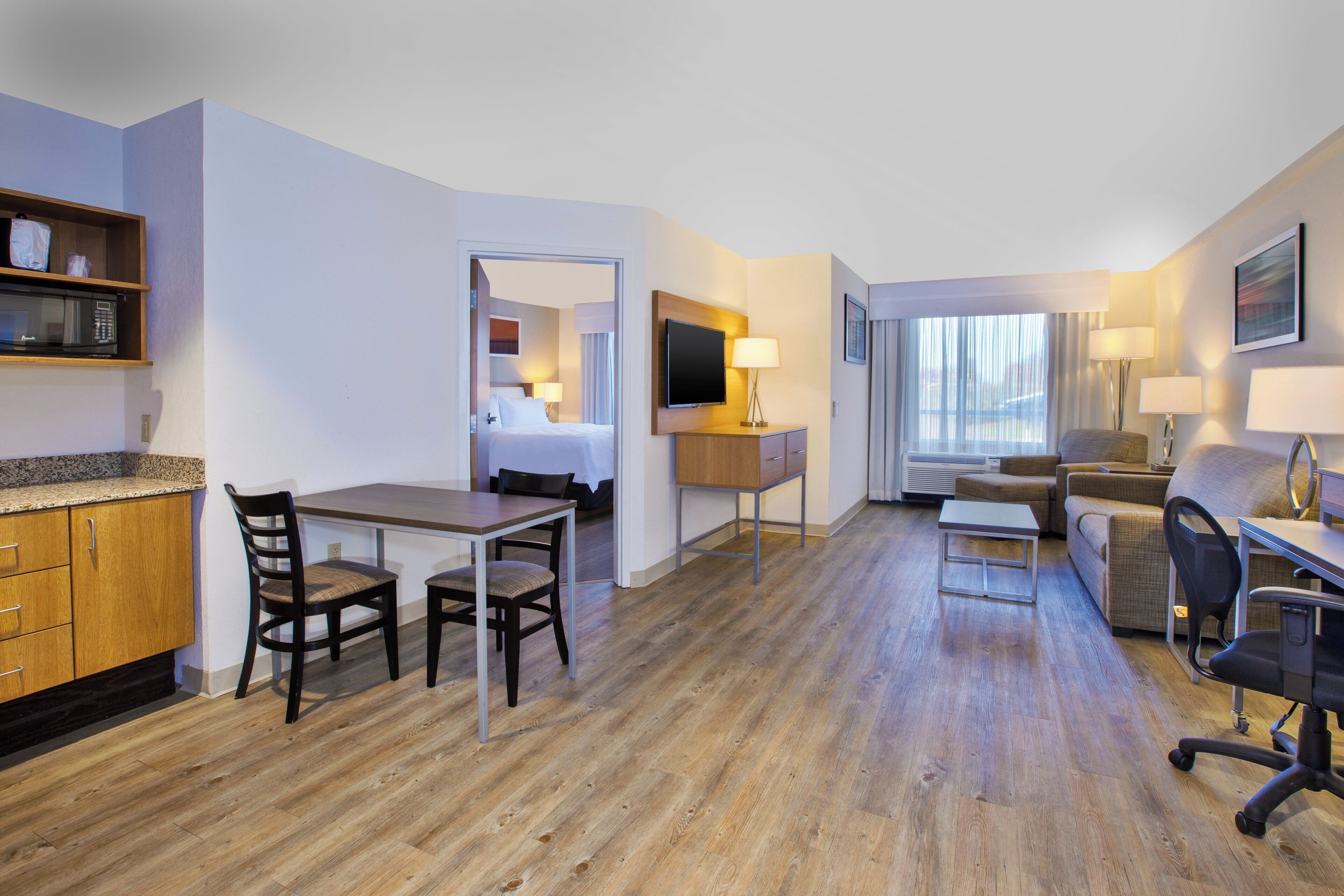 A hotel room with a wooden floor, a dining table with chairs, a sofa, a TV, a desk with a lamp, and a window with curtains.