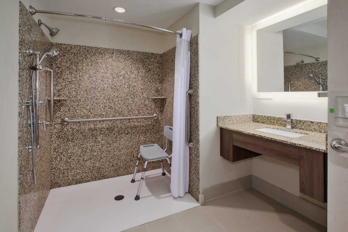A modern bathroom with a granite shower wall, a grab bar, a wheelchair-accessible bench, a sink with a wooden vanity, and a large mirror.