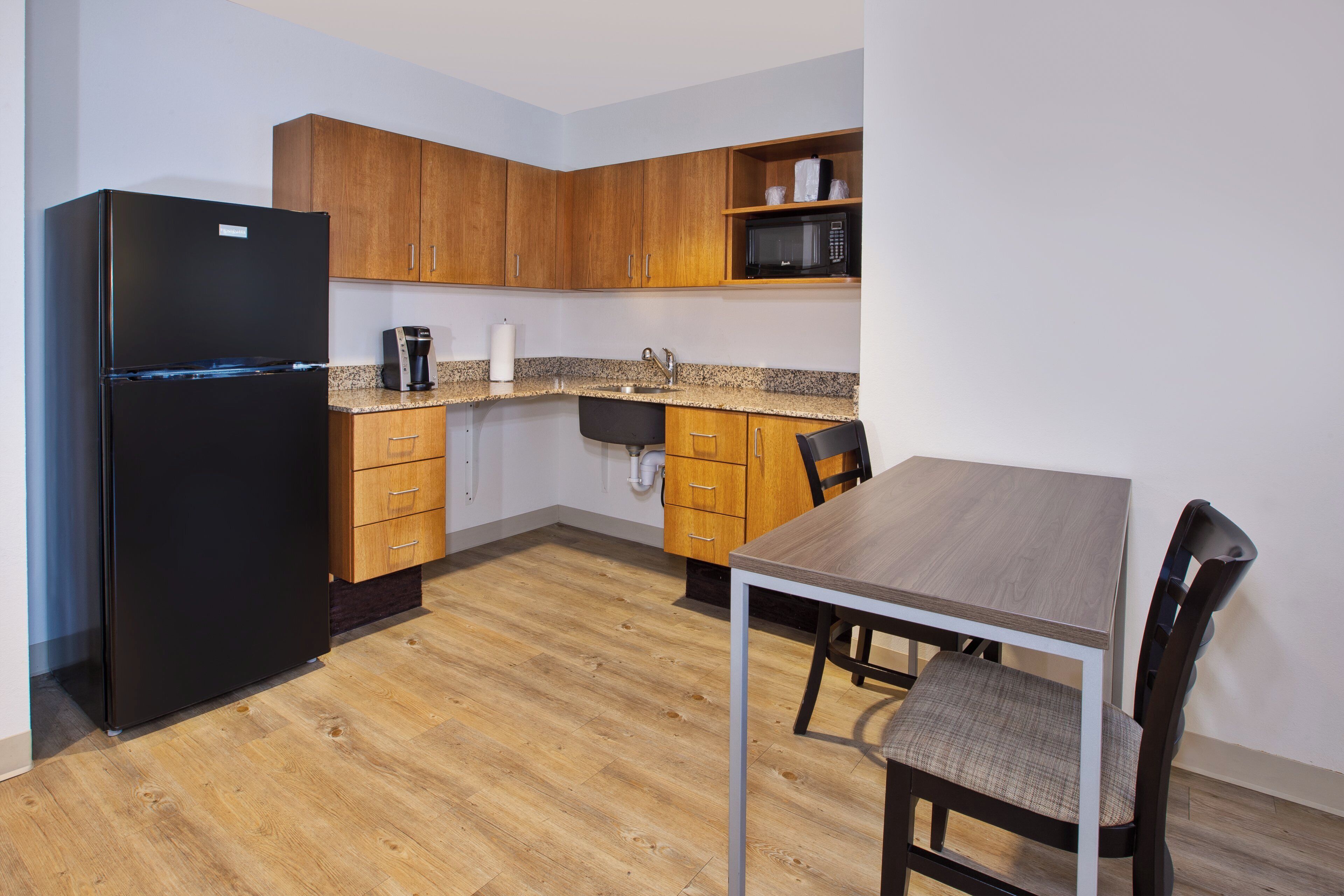 A compact kitchen with wooden cabinets, a black refrigerator, a coffee maker, and a dining table with chairs.