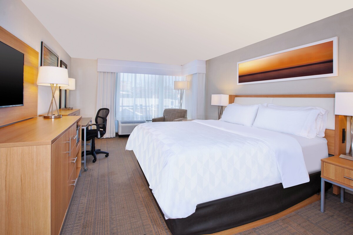A hotel room with a bed, desk, chair, and a large window.