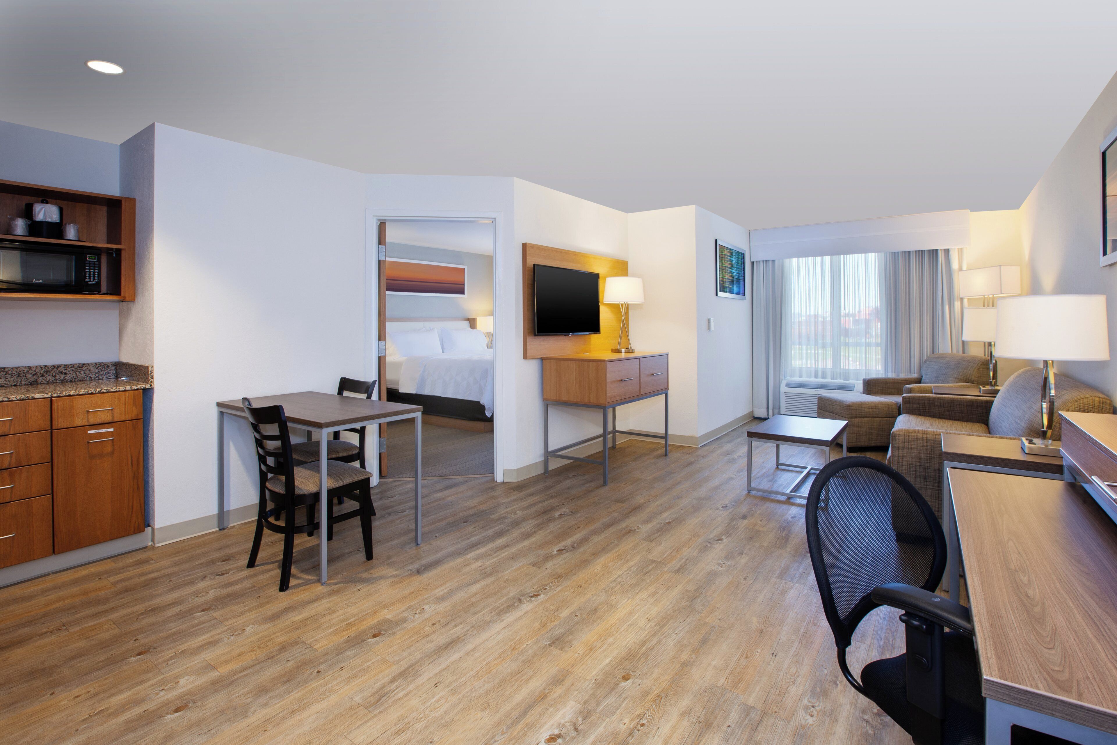 Habitación de hotel con kitchenette, zona de comedor, sala de estar con sofá y televisor, y un ventanal con cortinas.