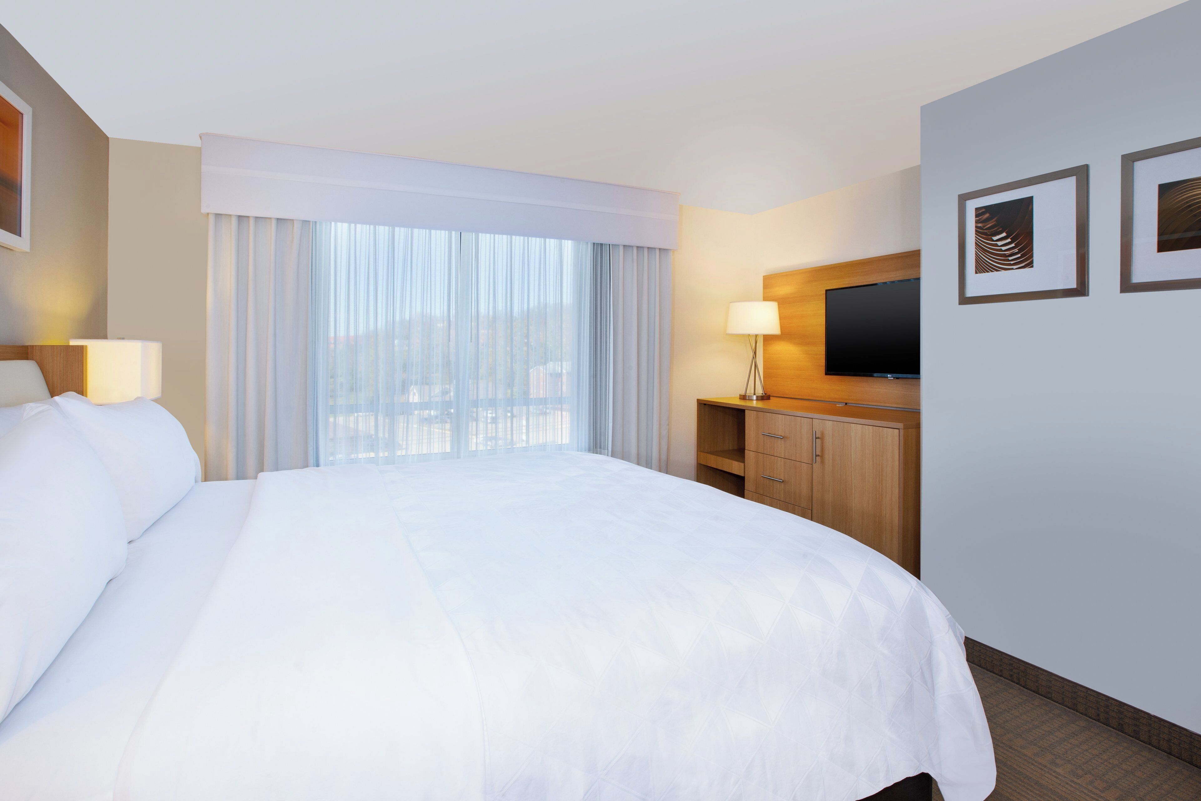 A hotel room with a large bed, a TV mounted on the wall, and a window with curtains.