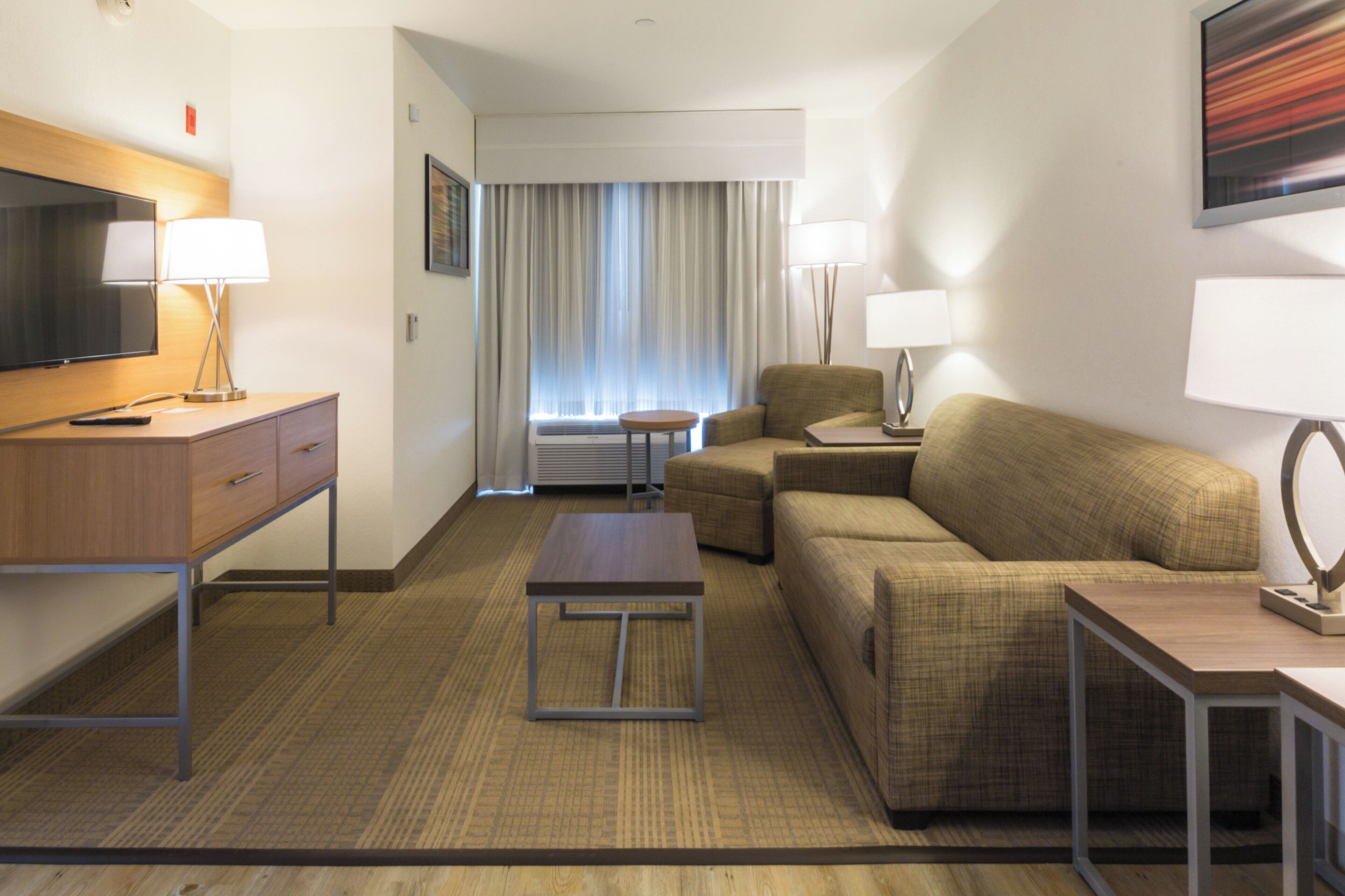 A hotel room with a television, a desk, a sofa, a small table, and a window with curtains.