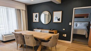 A modern hotel room with a wooden dining table, four chairs, a large mirror, and a bed with a white comforter.