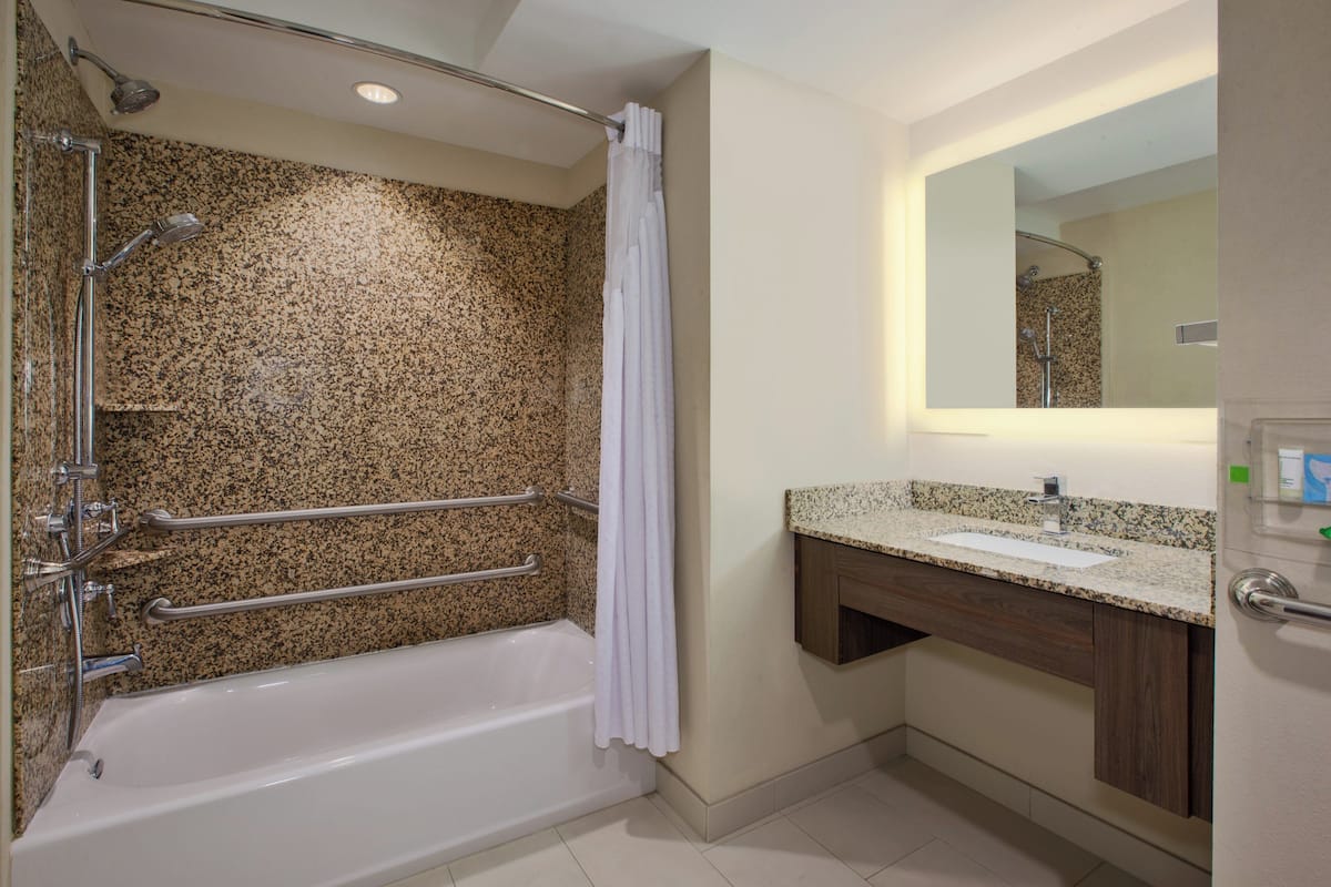 A bathroom with a granite shower wall, a white bathtub, a vanity with a sink, and a mirror above the sink.