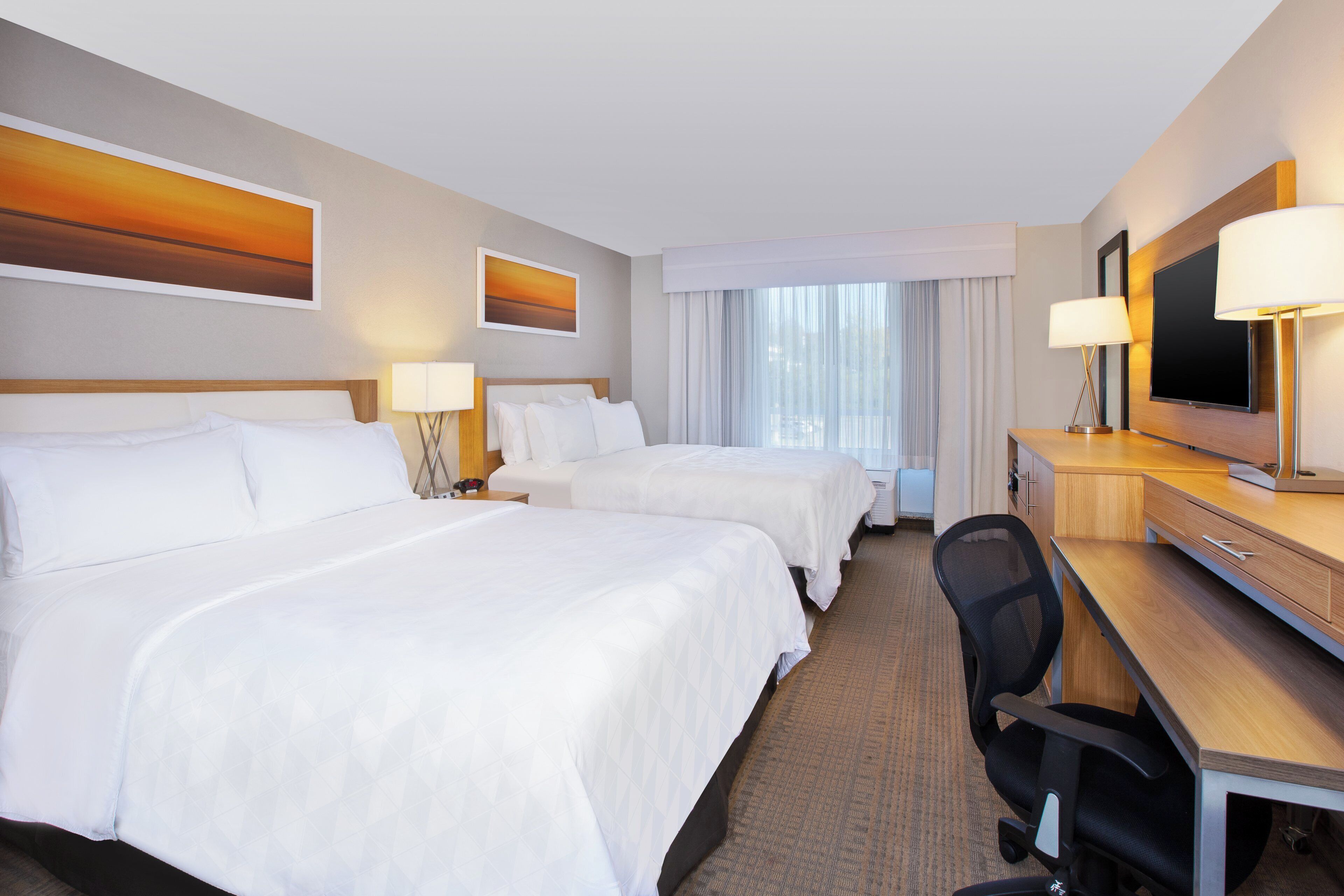 A hotel room with two beds, a desk, a TV, and a large window with curtains.