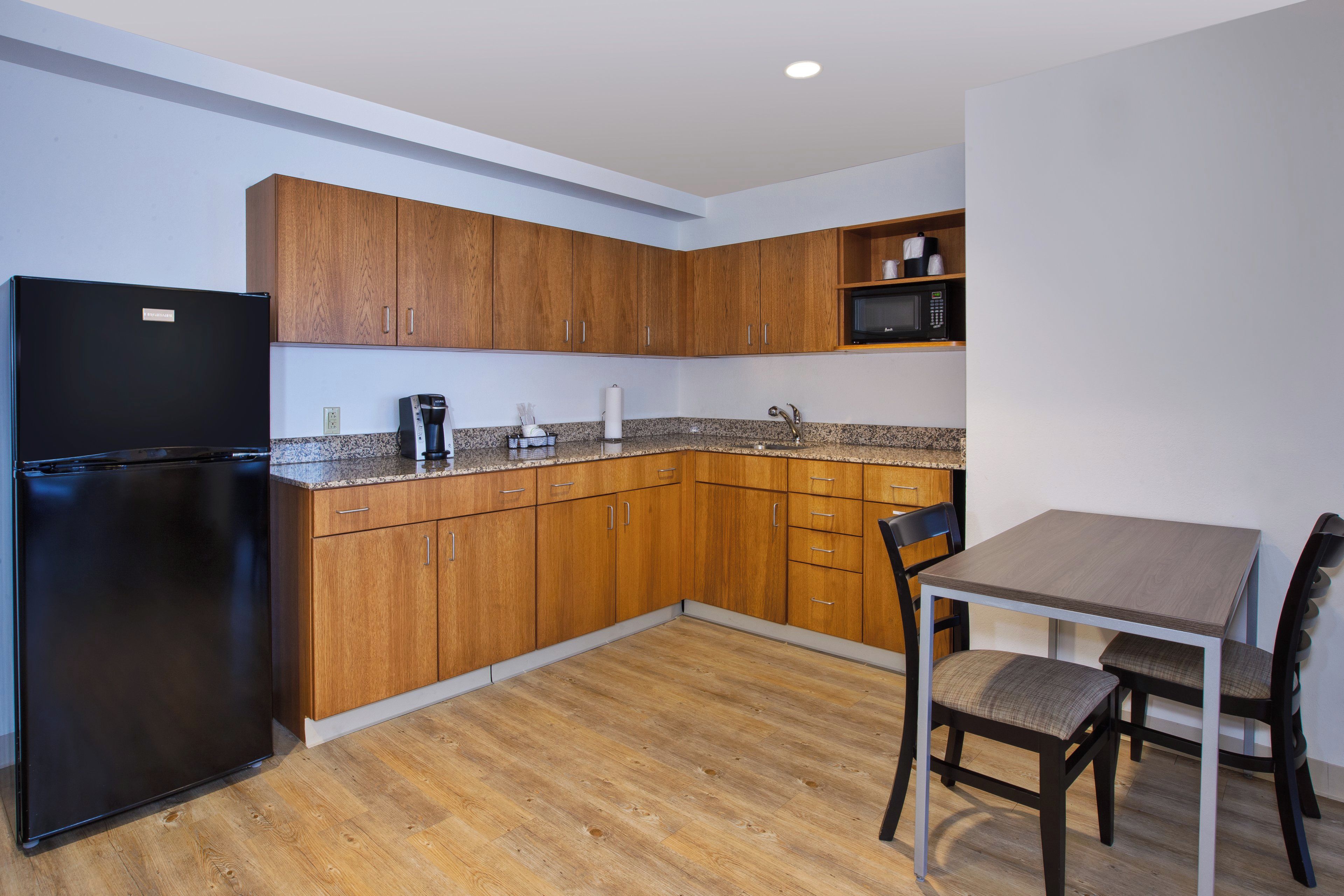 A compact kitchen with wooden cabinets, a black refrigerator, a microwave, and a dining table with chairs.