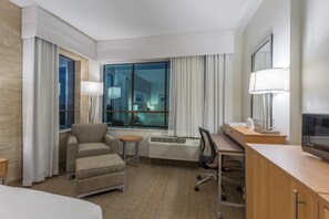 Een hotelkamer met een bed, een bureau met stoel, een bank, een klein rond tafeltje, een televisie en een groot raam met gordijnen.