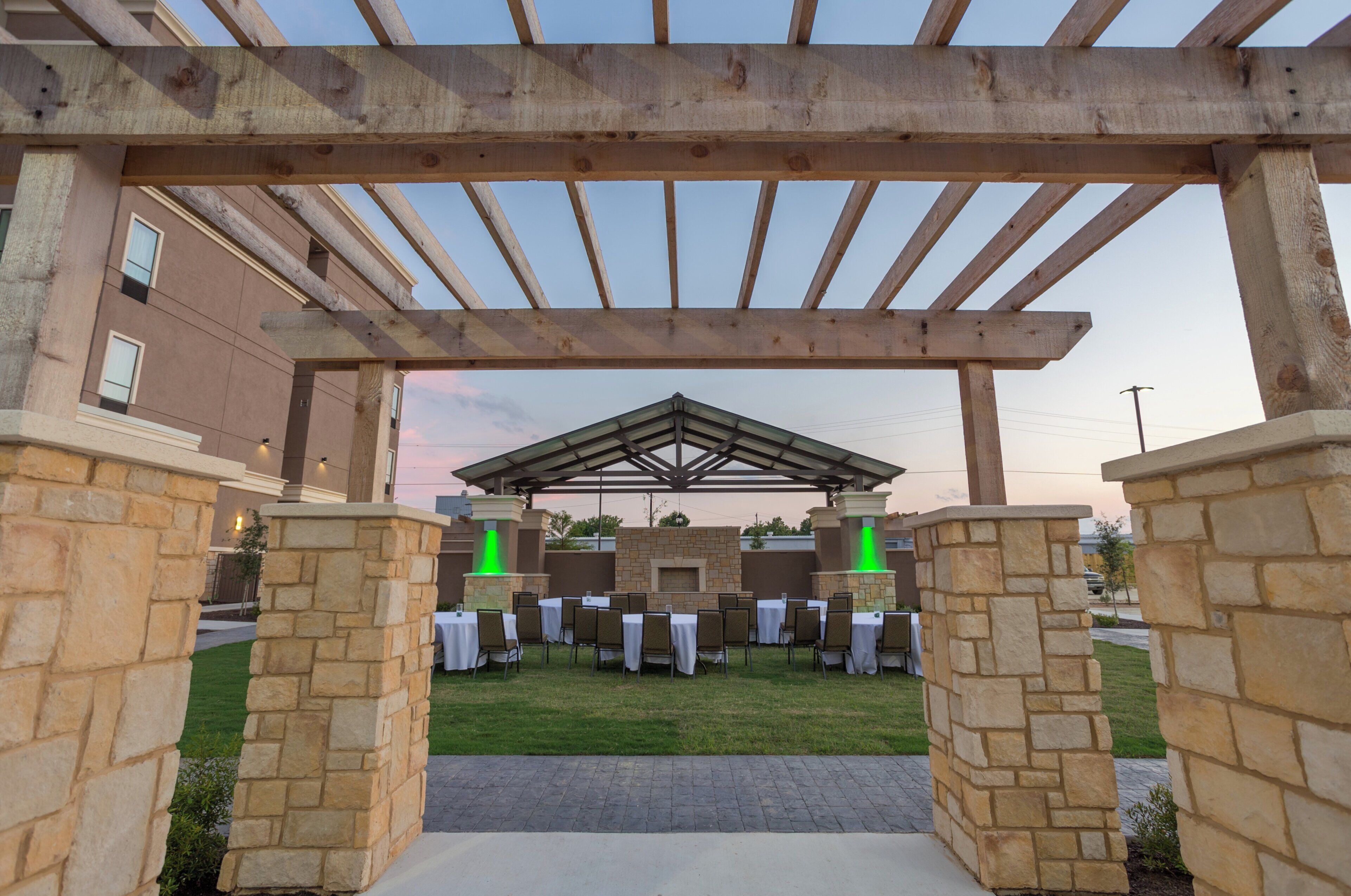 An outdoor patio area with stone pillars, tables, and chairs, under a wooden pergola structure.
