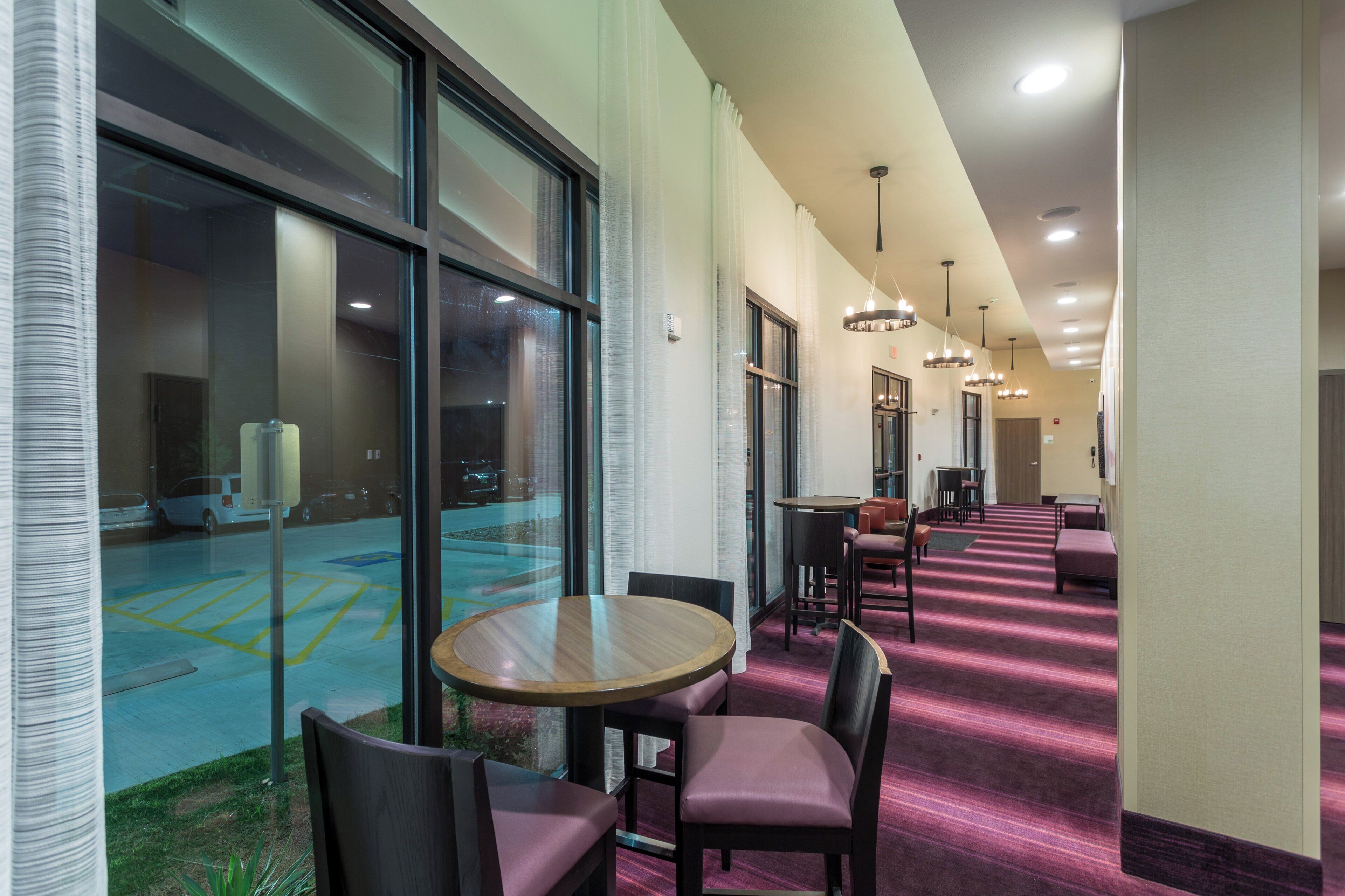 A hotel corridor with a carpeted floor, a round wooden table with chairs, and a view of a parking lot through large windows.