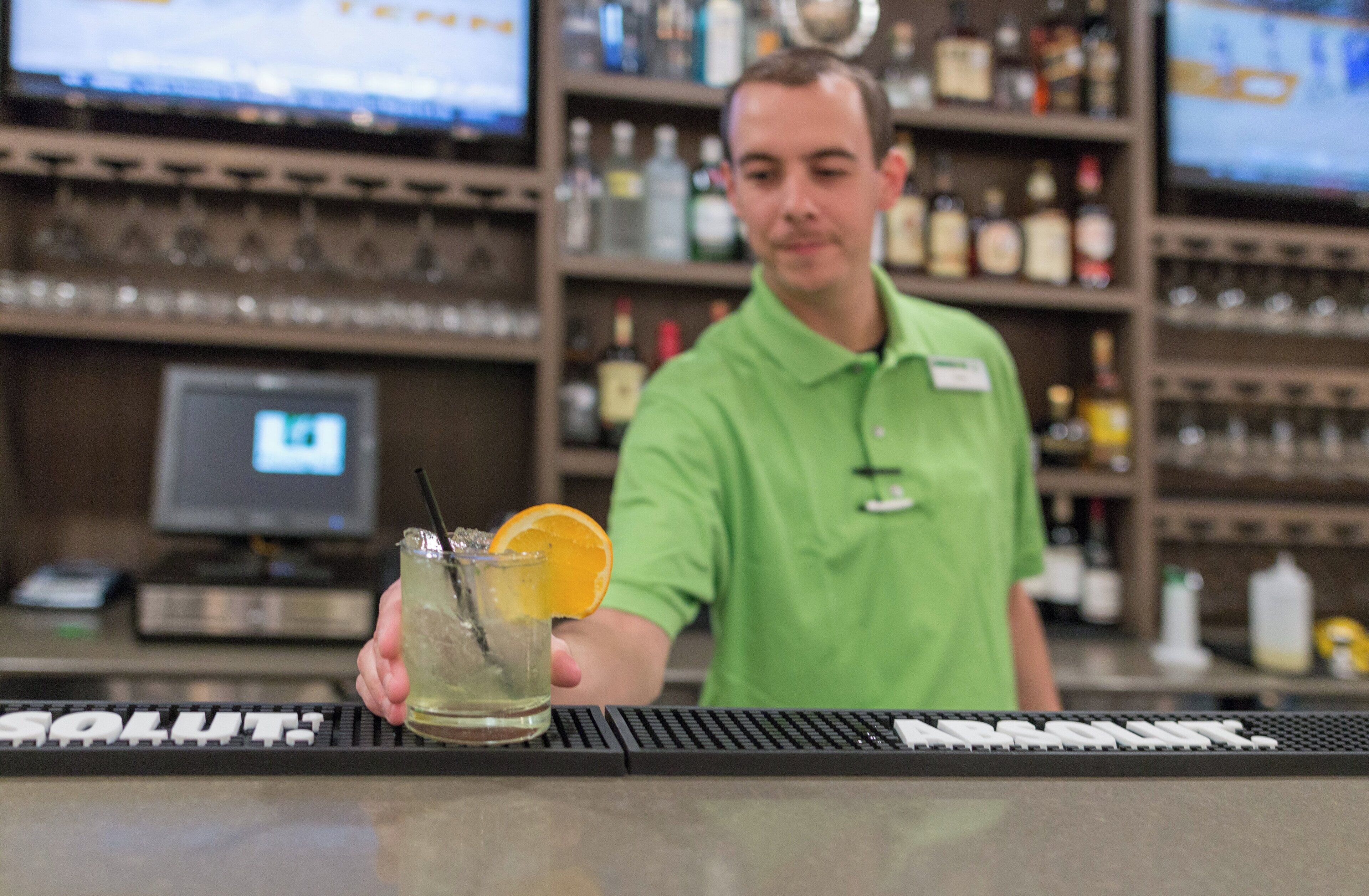 A bartender holding a drink with an orange slice, behind a bar with shelves of bottles and a computer monitor.