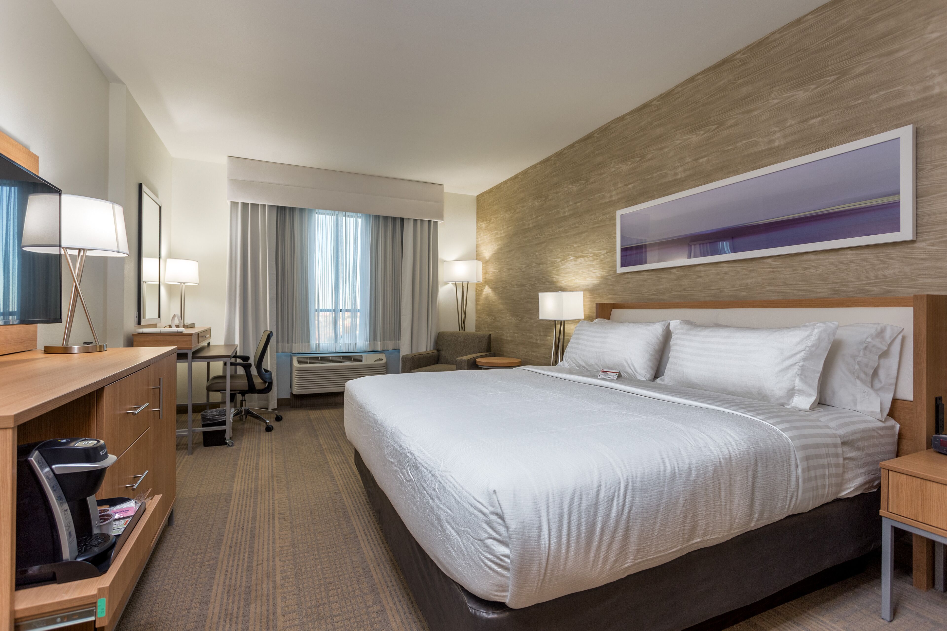 Habitación de hotel con una cama grande, un escritorio con silla, una lámpara de escritorio, una lámpara de pie, un televisor de pantalla plana y un ventanal con cortinas.