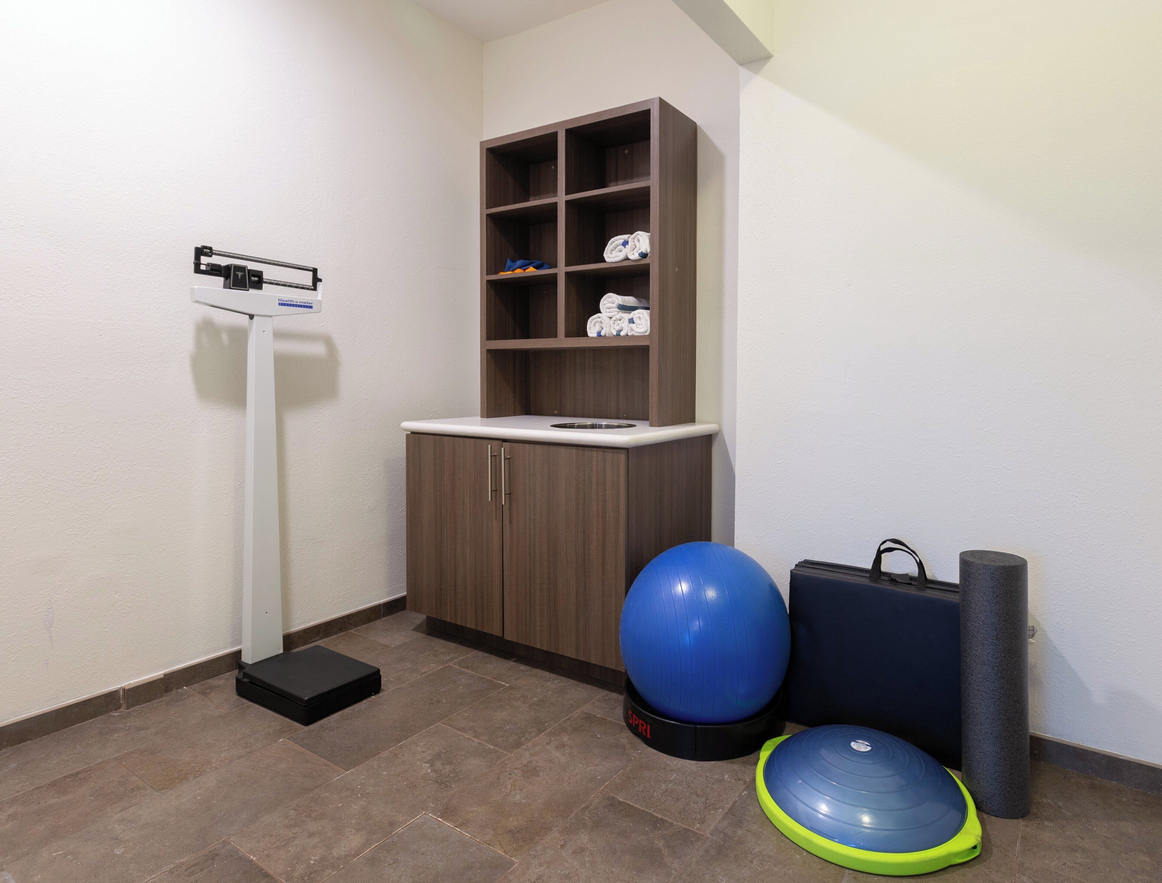 A room with a standing scale, a blue exercise ball, a yoga mat, and a cabinet with shelves.