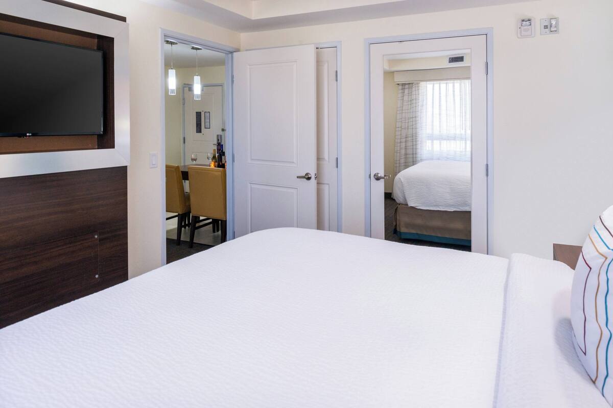 A hotel room with a large bed, a TV mounted on the wall, and two doors leading to a bathroom and a sitting area.