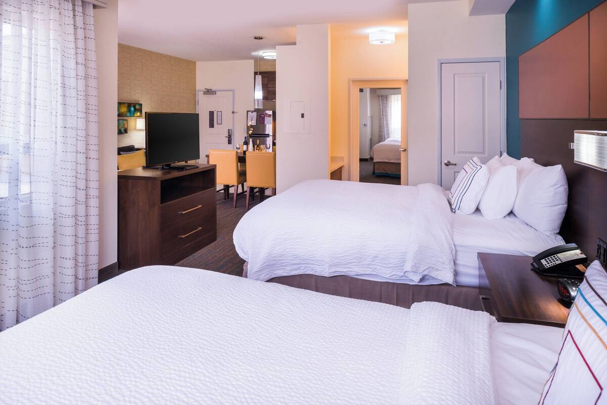 A hotel room with two beds, a desk with a TV, and a kitchenette in the background.
