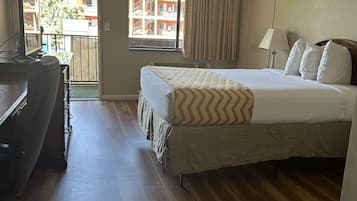 A hotel room with a bed, a desk, a chair, a window with curtains, and a view of a building outside.