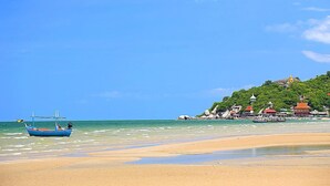 A beach with a boat, clear blue sky, and buildings on a hillside. - Chelona Khaotao Hua Hin E702 (Hua Hin)