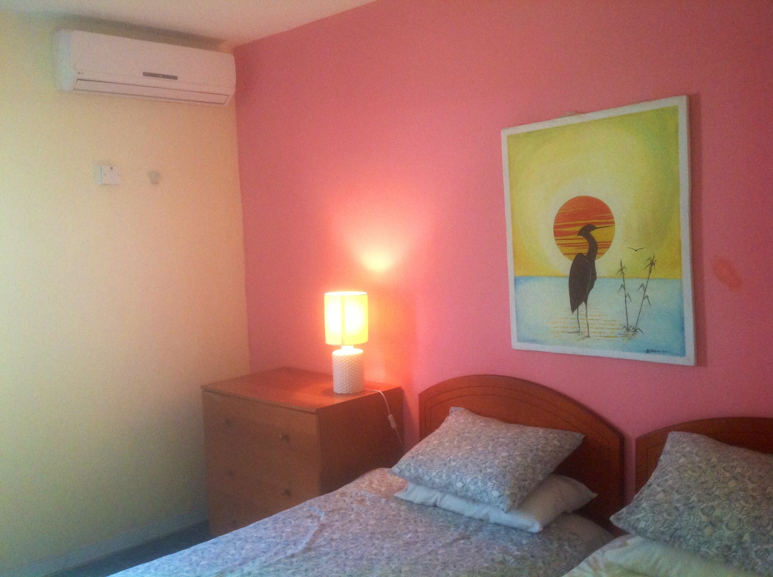 Quarto com cama, mesa de cabeceira com luminária, ar-condicionado na parede e quadro de um pássaro.