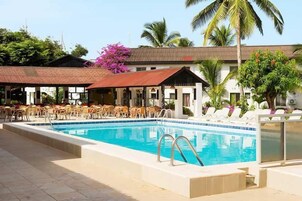 Una piscina con sillas de descanso y sombrillas, rodeada de palmeras y un edificio con techo de tejas.