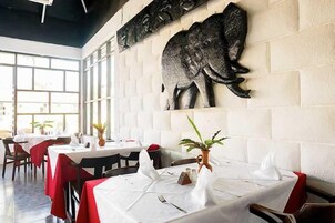 Un restaurante con paredes blancas, una gran escultura de elefante y mesas dispuestas con manteles blancos y servilletas rojas.
